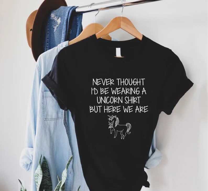 May include: Black t-shirt with white text that reads "Never thought I'd be wearing a unicorn shirt but here we are." A white outline of a unicorn is below the text.