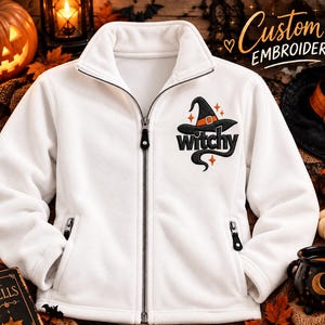 May include: A white fleece zip-up jacket featuring an embroidered design on the left chest. The embroidery depicts a black witch hat with an orange band, the word "witchy" in black script, and small orange star accents. The jacket has a high collar.