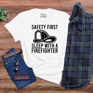 May include: White t-shirt with black text that reads "Safety First Sleep with a Firefighter" and a black silhouette of a firefighter's helmet.