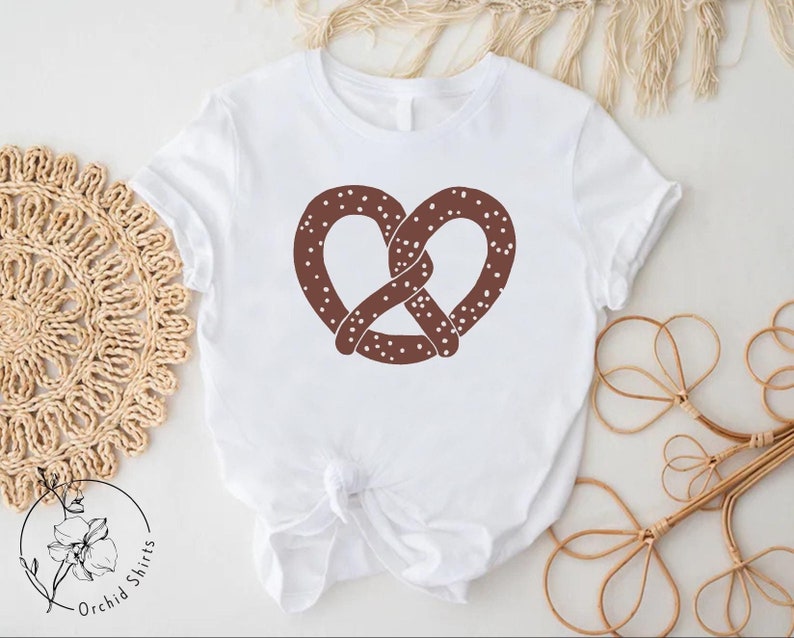 Pu&ograve; includere: Maglietta bianca con un design a forma di pretzel marrone. Il pretzel ha dei puntini bianchi. La maglietta &egrave; annodata in basso.