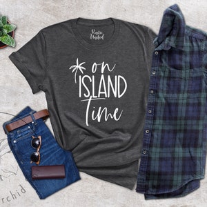 May include: A dark gray t-shirt with the text "on island time" in white lettering. The shirt is laid flat on a gray background with a pair of blue jeans, a brown leather belt, sunglasses, and a brown leather wallet.