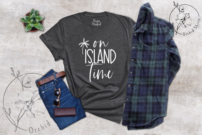 May include: A dark gray t-shirt with the text "on island time" in white lettering. The shirt is laid flat on a gray background with a pair of blue jeans, a brown leather belt, sunglasses, and a brown leather wallet.