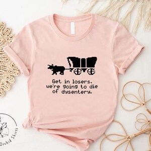Pu&ograve; includere: T-shirt rosa chiaro con grafica nera di un carro coperto trainato da un animale. Il testo "Get in losers, we're going to die of dysentery" &egrave; sotto. La maglietta ha maniche corte e girocollo.