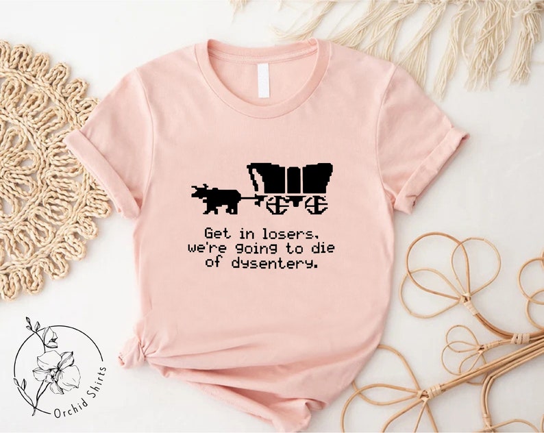 Pu&ograve; includere: T-shirt rosa chiaro con grafica nera di un carro coperto trainato da un animale. Il testo "Get in losers, we're going to die of dysentery" &egrave; sotto. La maglietta ha maniche corte e girocollo.