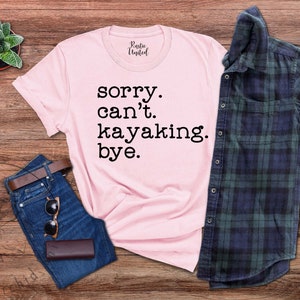 May include: Pink t-shirt with the text "sorry. can't. kayaking. bye." on a wooden background with a plaid shirt, jeans, and a potted plant.
