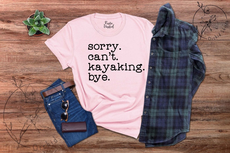 May include: Pink t-shirt with the text "sorry. can't. kayaking. bye." on a wooden background with a plaid shirt, jeans, and a potted plant.
