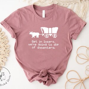 Pu&ograve; includere: T-shirt malva con un disegno pixelato bianco di un carro e buoi. Il testo "Get in losers, we're going to die of dysentery" &egrave; stampato sotto la grafica. La maglietta &egrave; di Orchid Shirts.