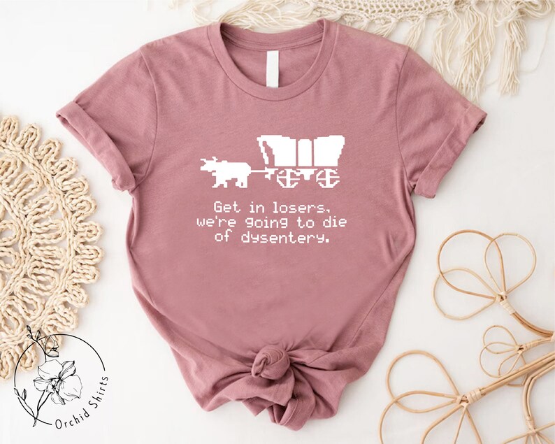 Pu&ograve; includere: T-shirt malva con un disegno pixelato bianco di un carro e buoi. Il testo "Get in losers, we're going to die of dysentery" &egrave; stampato sotto la grafica. La maglietta &egrave; di Orchid Shirts.
