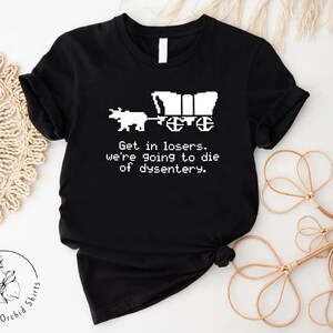 Pu&ograve; includere: T-shirt nero con un disegno pixelato di un carro e buoi. Il testo recita: "Get in losers, we're going to die of dysentery." La maglietta &egrave; di Orchid Shirts.