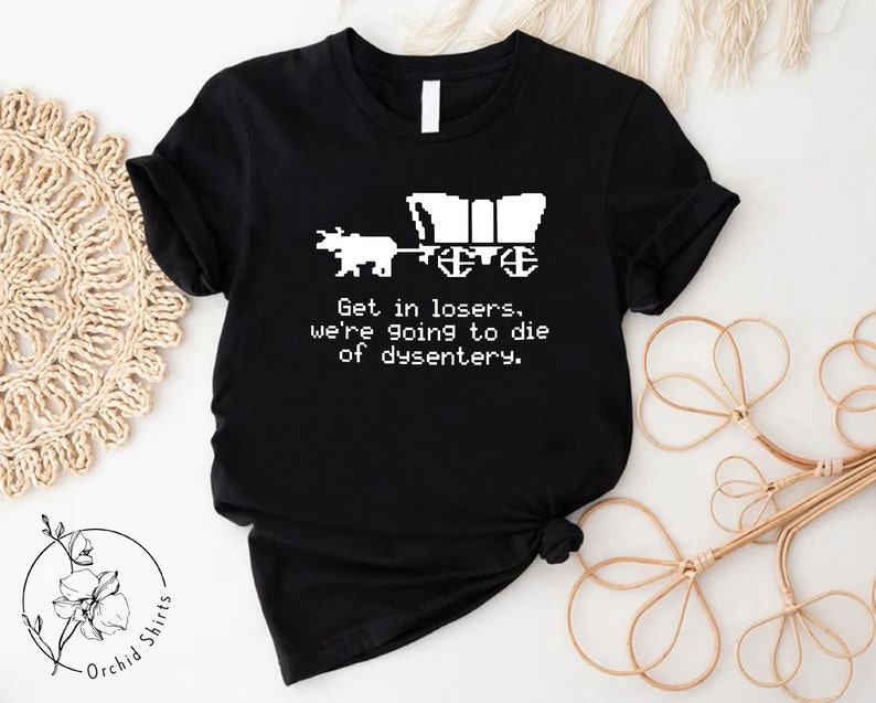 Pu&ograve; includere: T-shirt nero con un disegno pixelato di un carro e buoi. Il testo recita: "Get in losers, we're going to die of dysentery." La maglietta &egrave; di Orchid Shirts.