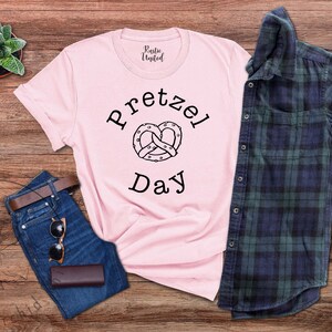 Op de afbeelding: Een roze T-shirt met een zwarte afbeelding van een pretzel en de tekst "Pretzel Day". Het shirt ligt op een houten oppervlak met een spijkerbroek, een zonnebril en een geruit overhemd.