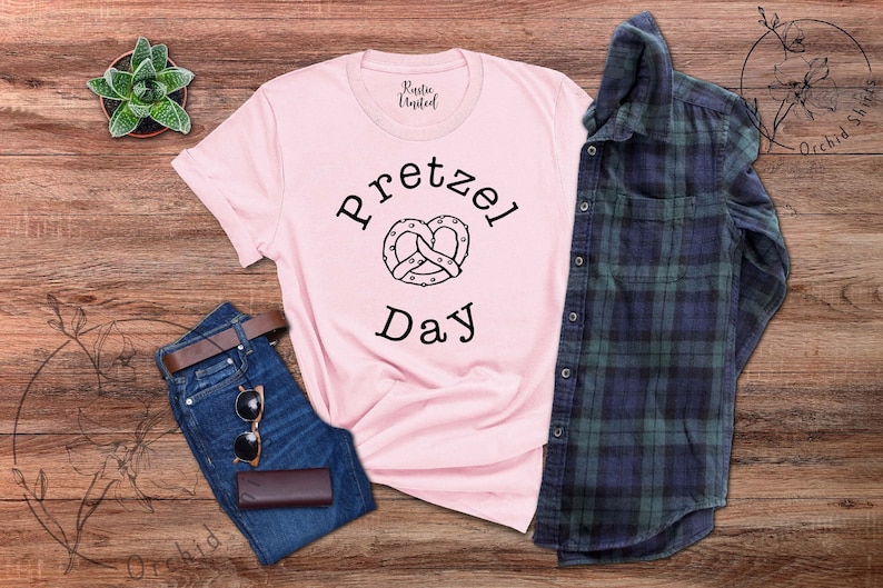 Op de afbeelding: Een roze T-shirt met een zwarte afbeelding van een pretzel en de tekst "Pretzel Day". Het shirt ligt op een houten oppervlak met een spijkerbroek, een zonnebril en een geruit overhemd.