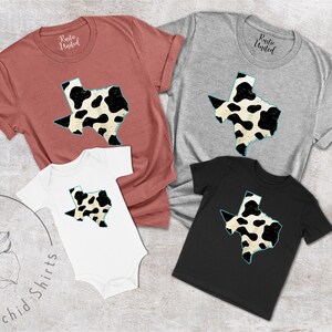 May include: A set of four shirts featuring a black and white cow print design of the state of Texas. The shirts are in various sizes and colors, including red, gray, white, and black.