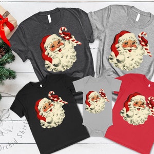 May include: A set of five t-shirts featuring a vintage illustration of Santa Claus holding a candy cane. The shirts are in various colors, including black, red, gray, and heather gray.