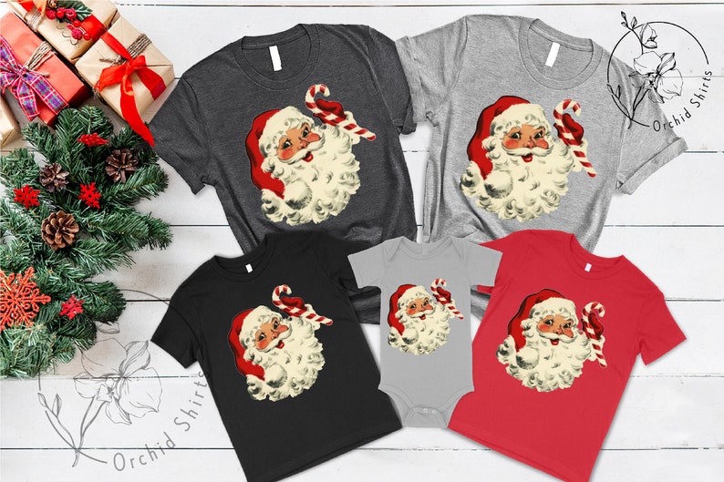 May include: A set of five t-shirts featuring a vintage illustration of Santa Claus holding a candy cane. The shirts are in various colors, including black, red, gray, and heather gray.