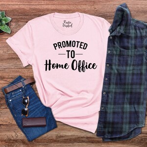 May include: A light pink t-shirt with the text "PROMOTED TO Home Office" in black. The shirt is laid out with a pair of blue jeans, a brown belt, sunglasses, a small brown wallet, a green and white succulent, and a blue and green plaid shirt.