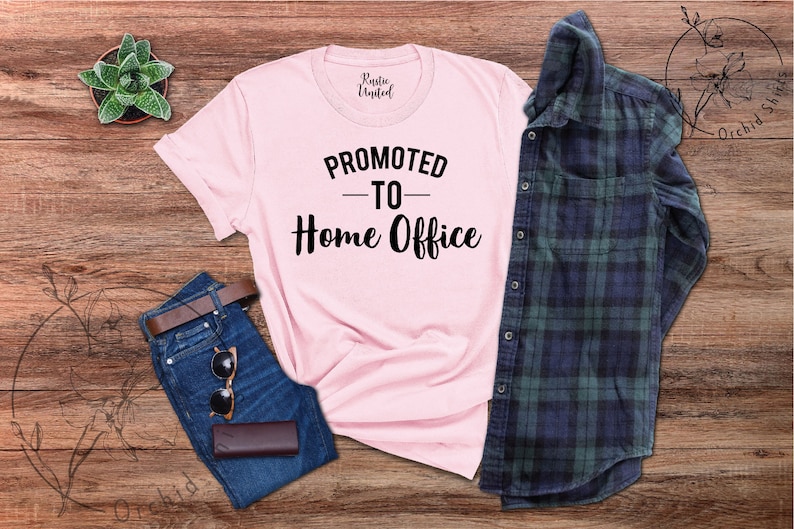 May include: A light pink t-shirt with the text "PROMOTED TO Home Office" in black. The shirt is laid out with a pair of blue jeans, a brown belt, sunglasses, a small brown wallet, a green and white succulent, and a blue and green plaid shirt.