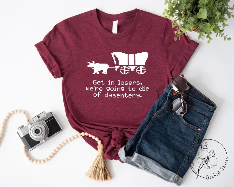 Pu&ograve; includere: T-shirt bordeaux con grafica bianca di un carro coperto trainato da buoi. Il testo "Get in losers, we're going to die of dysentery" &egrave; stampato sotto. La maglietta &egrave; abbinata a pantaloncini di jeans e occhiali da sole.