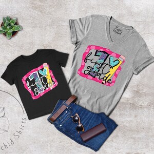 May include: Two t-shirts with the text "I love first grade" in a colourful design. The shirts are black and grey and have a heart and pencil graphic.