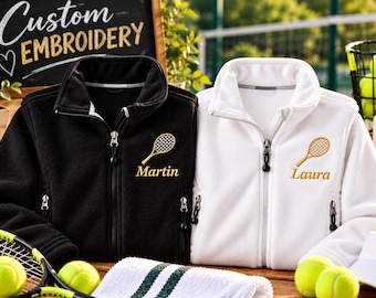 Op de afbeelding: Twee fleece jassen, één zwart en één wit, elk voorzien van een geborduurd gouden tennisracket en een naam. De zwarte jas is geborduurd met Martin en de witte jas met Laura. Beide jassen hebben een ritssluiting over de gehele lengte.