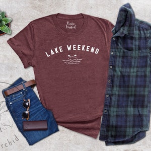 以下が含まれることがあります： 白い「LAKE WEEKEND」の文字とオールと波のグラフィックが入った、マルーン色のTシャツ。青いジーンズ、茶色のベルト、サングラス、財布も写っています。青と緑のチェック柄のシャツも含まれています。