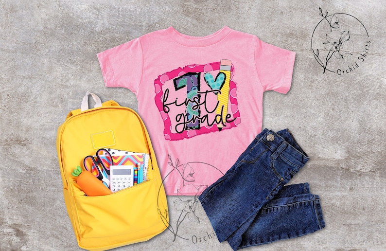 May include: A pink t-shirt with a colourful graphic that says "1st grade" with a pencil and a heart. The shirt is lying on a grey surface with a yellow backpack, a pair of blue jeans, and a white flower.