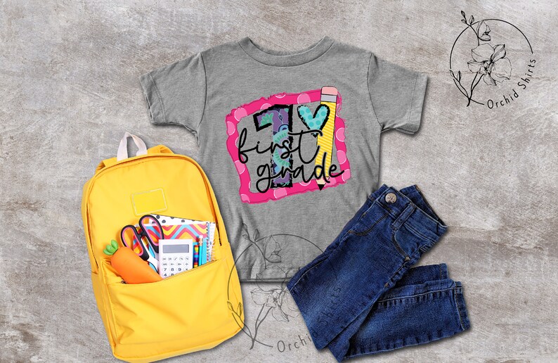 May include: A grey t-shirt with a pink and white graphic that says "1st grade" with a pencil and heart design. A yellow backpack with school supplies, a pair of blue jeans, and a grey background.