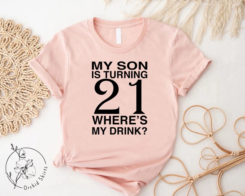 a pink shirt that says, my son is turning 21 where's my drink