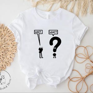 May include: White t-shirt with black text and illustrations. The text says "WAIT." and "WHAT?" with a question mark character above the word "WHAT?"