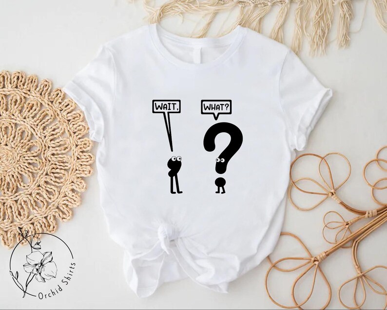 May include: White t-shirt with black text and illustrations. The text says "WAIT." and "WHAT?" with a question mark character above the word "WHAT?"