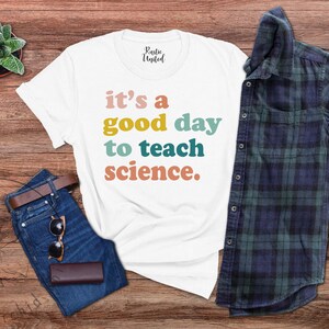 Op de afbeelding: Wit T-shirt met de tekst "it's a good day to teach science." in kleurrijke letters. Het shirt ligt op een houten oppervlak met een blauwe jeans, een zonnebril en een geruit overhemd.