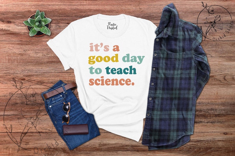 Op de afbeelding: Wit T-shirt met de tekst "it's a good day to teach science." in kleurrijke letters. Het shirt ligt op een houten oppervlak met een blauwe jeans, een zonnebril en een geruit overhemd.
