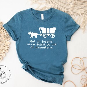 Pu&ograve; includere: T-shirt color teal con un disegno pixelato di un carro e buoi. Il testo recita: "Get in losers, we're going to die of dysentery." La maglietta &egrave; di Orchid Shirts.