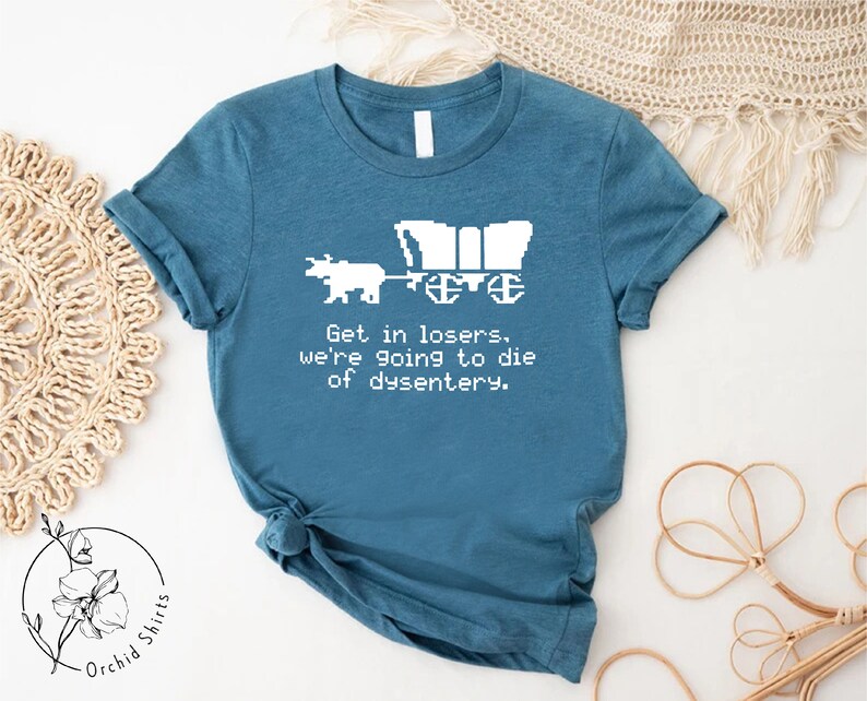 Pu&ograve; includere: T-shirt color teal con un disegno pixelato di un carro e buoi. Il testo recita: "Get in losers, we're going to die of dysentery." La maglietta &egrave; di Orchid Shirts.