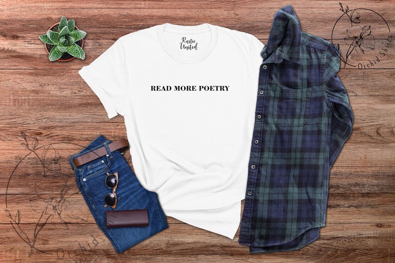 Op de afbeelding: Wit T-shirt met de tekst "READ MORE POETRY" in zwart gedrukt op de voorkant. Het shirt ligt plat op een houten oppervlak met een spijkerbroek, een zonnebril en een geruit overhemd.