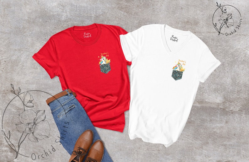 May include: Two t-shirts, one red and one white, with a pocket design featuring a unicorn. The pocket has a black and white pattern. The red shirt is crew neck, and the white shirt is a V-neck. Jeans and brown shoes are also in the image.