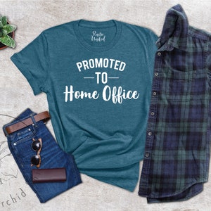 May include: Teal t-shirt with the text "PROMOTED TO - Home Office" in white. The shirt is paired with blue jeans, a brown belt, sunglasses, and a wallet. A plaid shirt and a small potted plant are also in the image.
