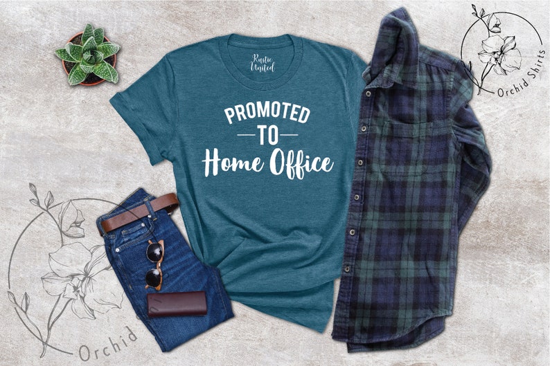 May include: Teal t-shirt with the text "PROMOTED TO - Home Office" in white. The shirt is paired with blue jeans, a brown belt, sunglasses, and a wallet. A plaid shirt and a small potted plant are also in the image.