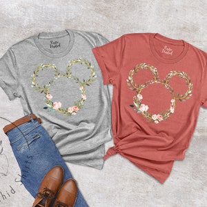 Peut inclure: Deux t-shirts, un gris et un corail, chacun avec un motif floral de Mickey Mouse. Le motif comprend un contour dor&eacute; avec des feuilles et des fleurs roses. Les chemises sont pr&eacute;sent&eacute;es avec un jean et des chaussures marron.