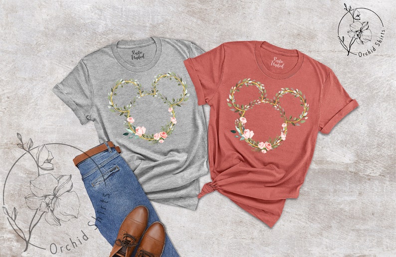 Peut inclure: Deux t-shirts, un gris et un corail, chacun avec un motif floral de Mickey Mouse. Le motif comprend un contour dor&eacute; avec des feuilles et des fleurs roses. Les chemises sont pr&eacute;sent&eacute;es avec un jean et des chaussures marron.