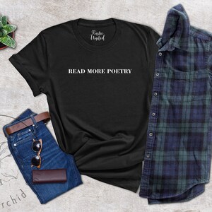 Op de afbeelding: Zwart T-shirt met de tekst "READ MORE POETRY" in witte letters. Het shirt ligt plat op een grijs, getextureerd oppervlak met een spijkerbroek, een bruine leren riem, een zonnebril en een kleine plant in een pot.