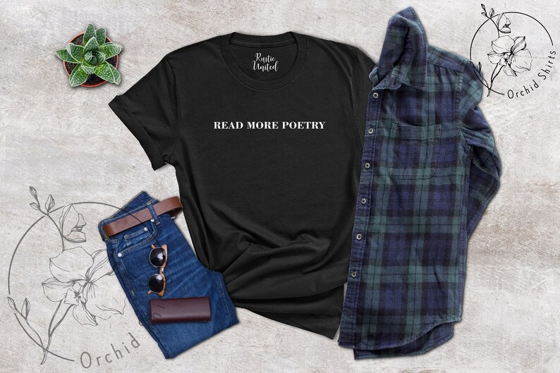 Op de afbeelding: Zwart T-shirt met de tekst "READ MORE POETRY" in witte letters. Het shirt ligt plat op een grijs, getextureerd oppervlak met een spijkerbroek, een bruine leren riem, een zonnebril en een kleine plant in een pot.