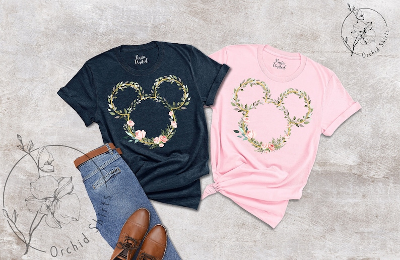 Peut inclure: Deux t-shirts, un bleu marine et un rose, chacun avec un motif floral de t&ecirc;te de Mickey Mouse. Le motif est compos&eacute; de feuilles vertes, d'accents dor&eacute;s et de fleurs roses. Les chemises sont pr&eacute;sent&eacute;es avec un jean et des chaussures marron.