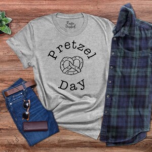 Op de afbeelding: Een grijs T-shirt met de tekst "Pretzel Day" en een zwart-witte illustratie van een pretzel. Het shirt ligt op een houten oppervlak met een spijkerbroek, een zonnebril en een geruit overhemd.