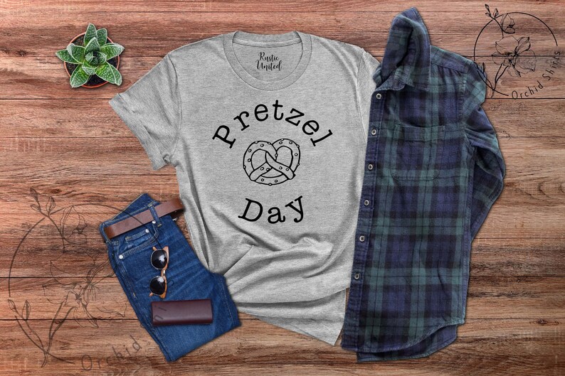 Op de afbeelding: Een grijs T-shirt met de tekst "Pretzel Day" en een zwart-witte illustratie van een pretzel. Het shirt ligt op een houten oppervlak met een spijkerbroek, een zonnebril en een geruit overhemd.