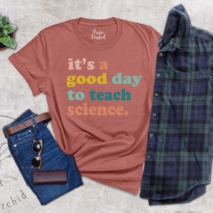 Op de afbeelding: Een roestkleurig T-shirt met de tekst "it's a good day to teach science." in kleurrijke letters. Het shirt ligt op een lichtgrijze achtergrond met een spijkerbroek, een bruine leren portemonnee, een zonnebril en een potplant.