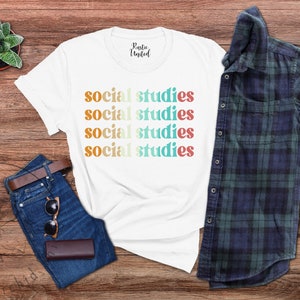May include: White t-shirt with the words "social studies" in a repeating design of orange, yellow, teal, and red. The shirt is next to a pair of blue jeans, sunglasses, a brown wallet, a brown belt, a small succulent plant, and a blue and green plaid shirt.
