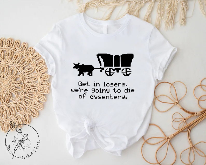 Pu&ograve; includere: T-shirt bianca con grafica pixelata nera di un carro coperto trainato da un bue. Il testo recita: "Get in losers, we're going to die of dysentery". La maglietta &egrave; di Orchid Shirts.