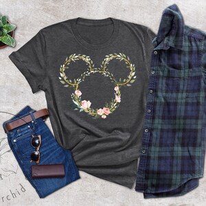 Peut inclure: T-shirt gris fonc&eacute; avec un motif floral de Mickey Mouse. Le motif comprend un contour dor&eacute; avec des feuilles vertes et des fleurs roses. L'image comprend &eacute;galement un jean bleu, une ceinture marron, des lunettes de soleil et une chemise &agrave; carreaux.