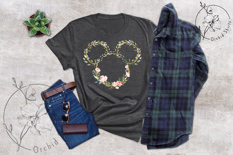Peut inclure: T-shirt gris fonc&eacute; avec un motif floral de Mickey Mouse. Le motif comprend un contour dor&eacute; avec des feuilles vertes et des fleurs roses. L'image comprend &eacute;galement un jean bleu, une ceinture marron, des lunettes de soleil et une chemise &agrave; carreaux.